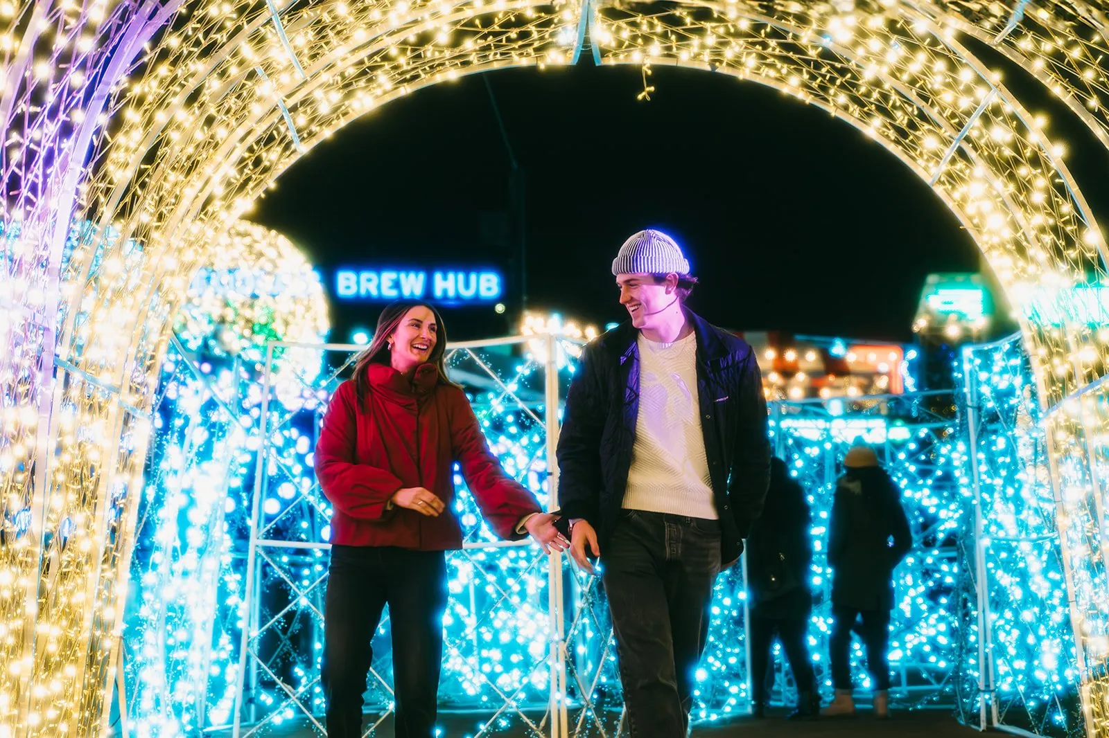 A couple holds hands under an arch of Christmas lights