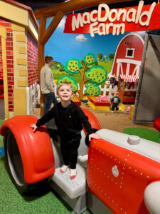 A boy plays on a large toy tractor. Sign in the background says MacDonald Farm.