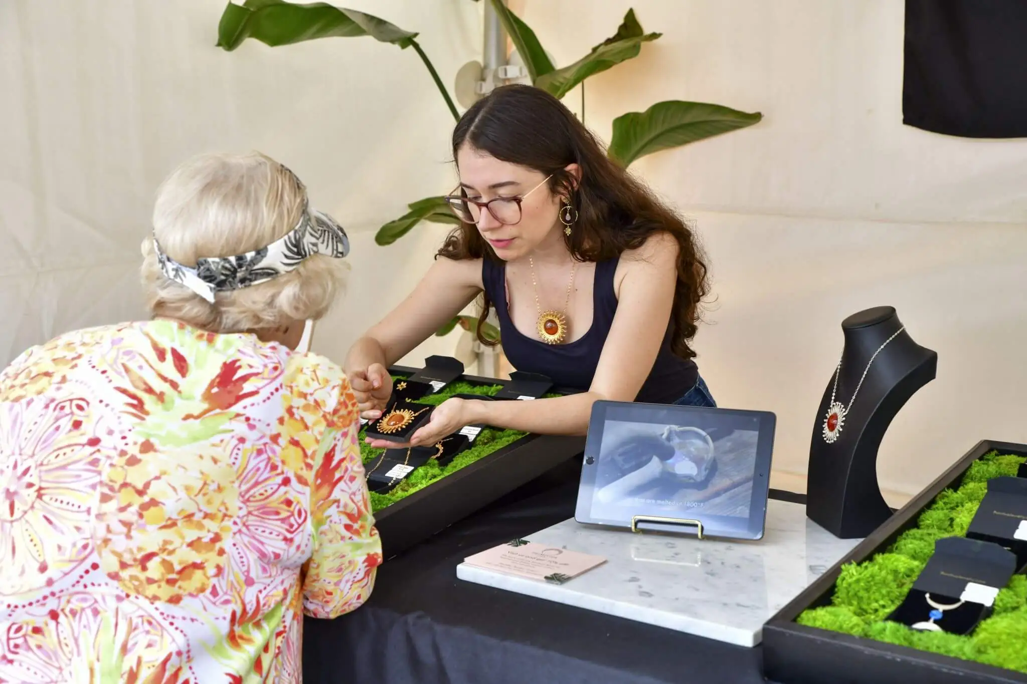 A woman shows her jewelry to a shopper at an art fair