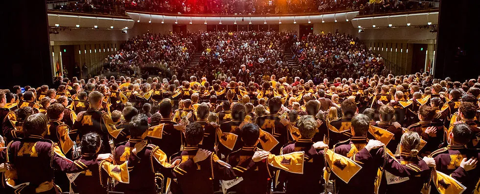 Rear view of a group of marching band performers with their arms wrapped around each other, looking out at a crowd