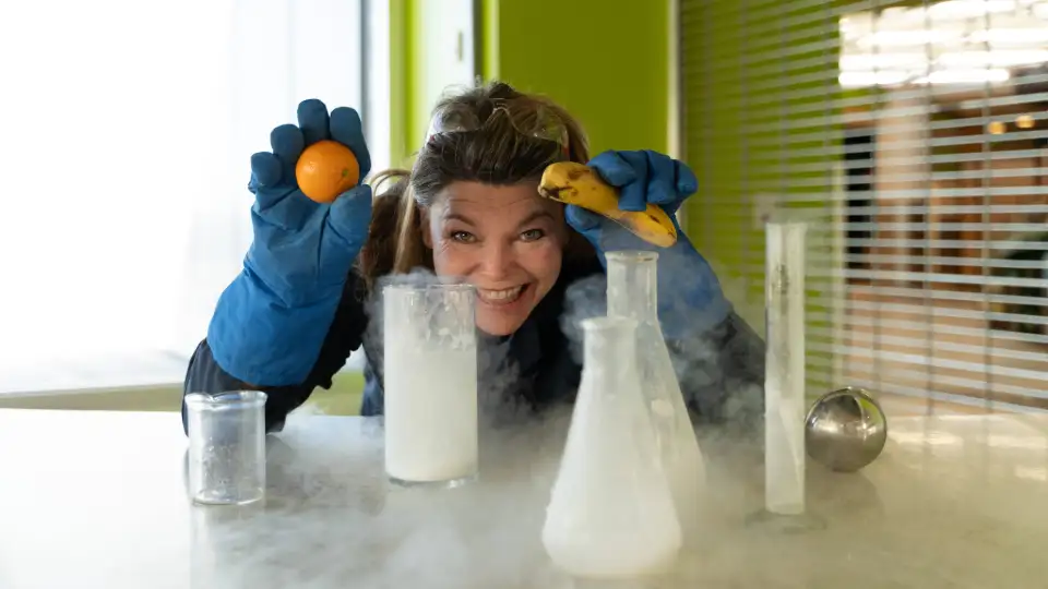 A smiling woman holds a banana and orange while looking out over an assortment of bubbling beakers